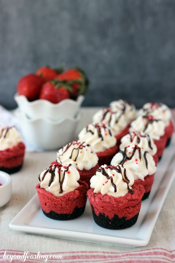 two lines of red velvet cream pie cookie cups on a white plate next to a bowl of strawberries