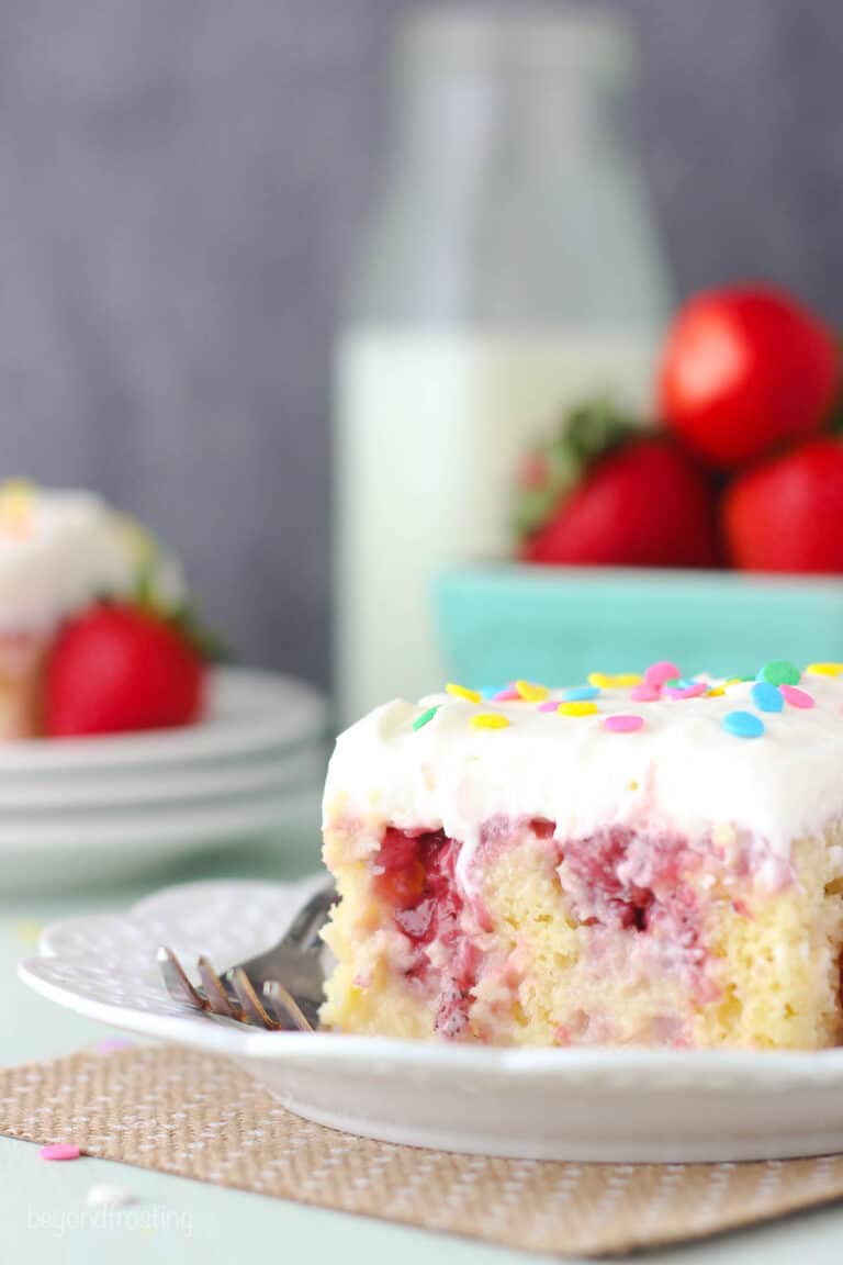 a slice of strawberry poke cake on a plate with a fork and strawberries in the background