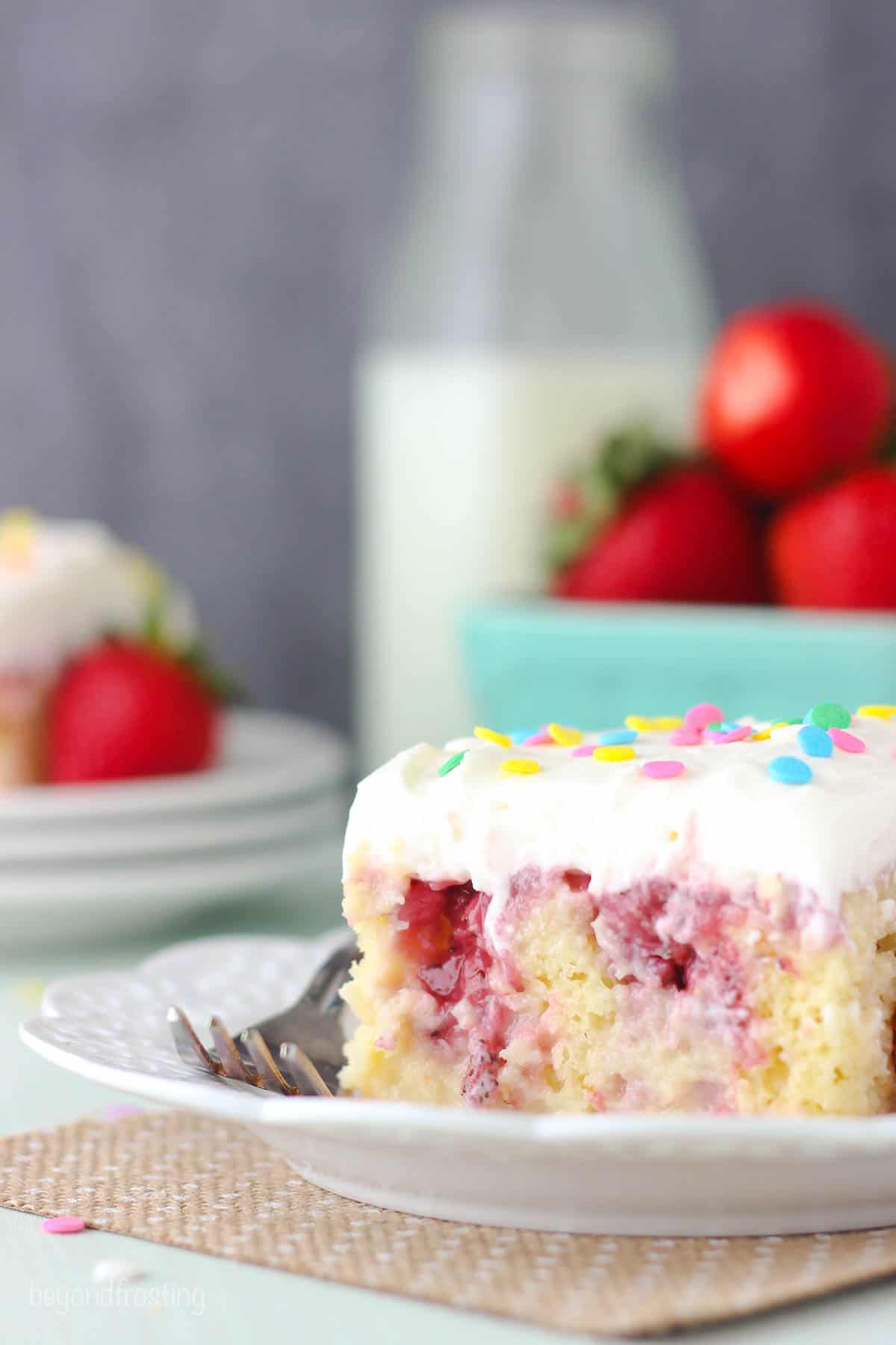 a slice of strawberry poke cake on a plate with a fork and strawberries in the background