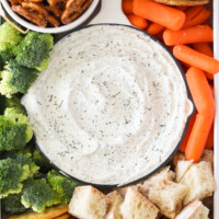 Close up of a bowl of dill dip with broccoli, carrots, bread and pretzels.