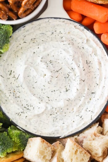 Close up of a bowl of dill dip with broccoli, carrots, bread and pretzels.