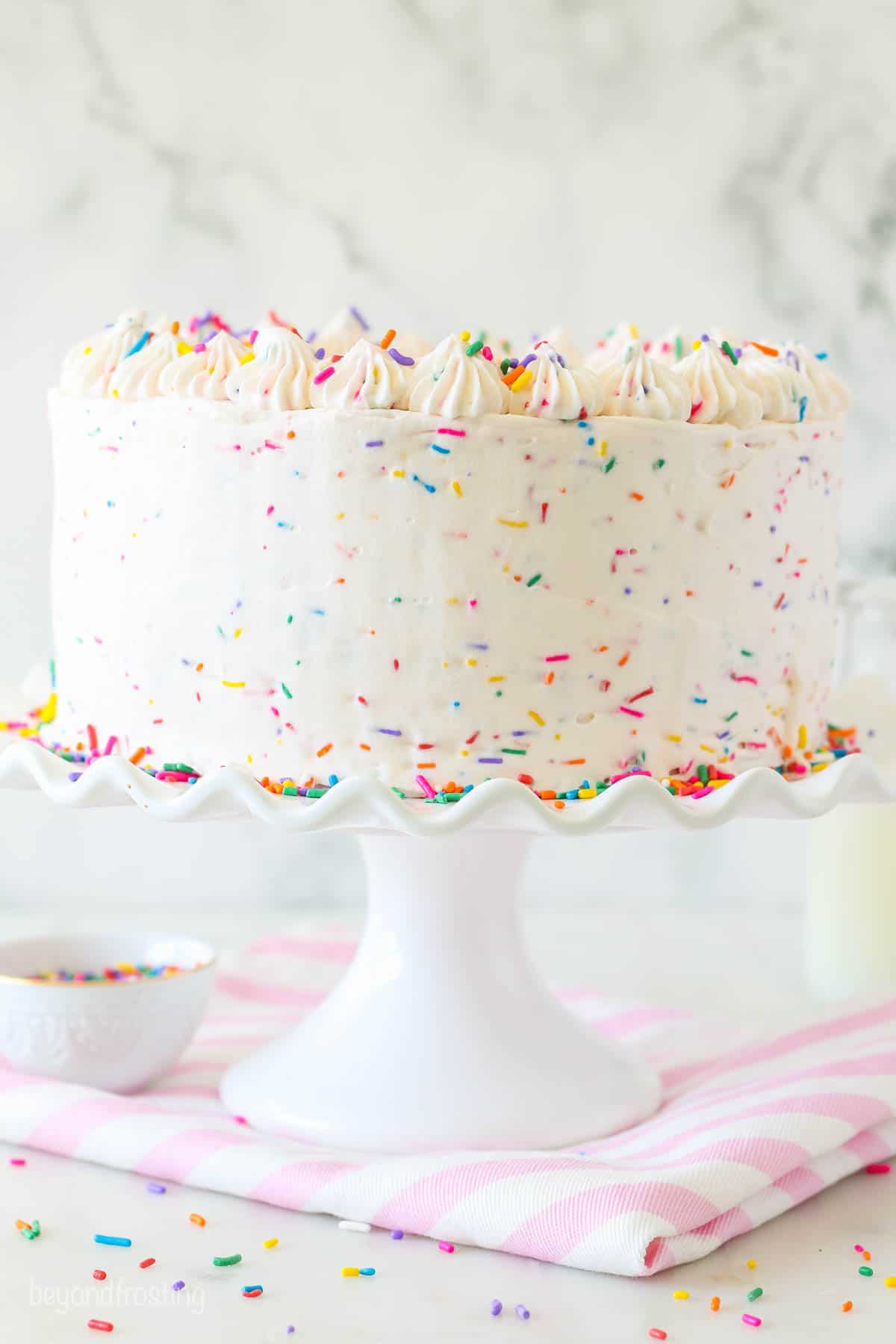 A cake decorated with sprinkles buttercream on a white cake plate with a pink striped napkin