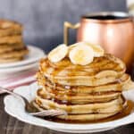 a large stack of pancakes on a plate topped with banana slices and doused in maple syrup