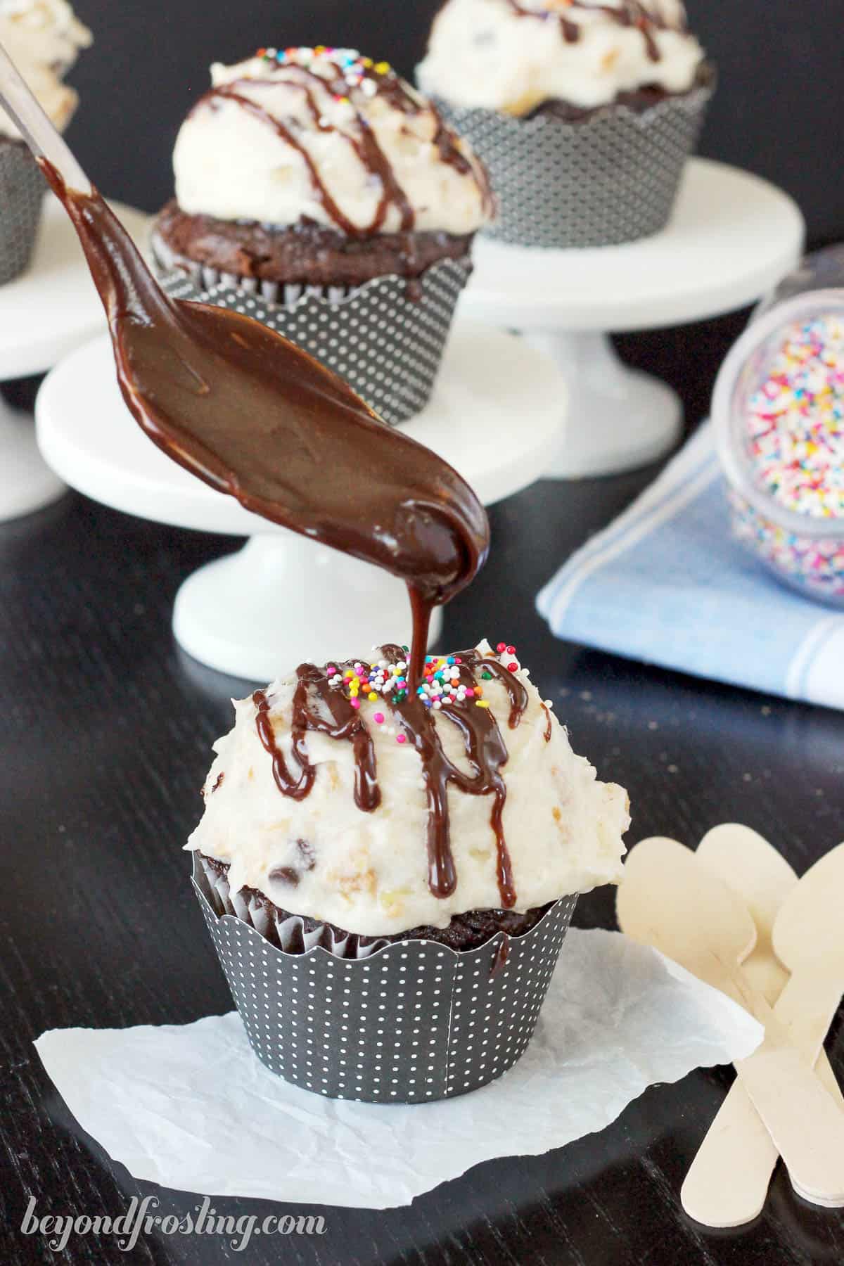 hot fudge being drizzled over a frosted cupcake topped with sprinkles