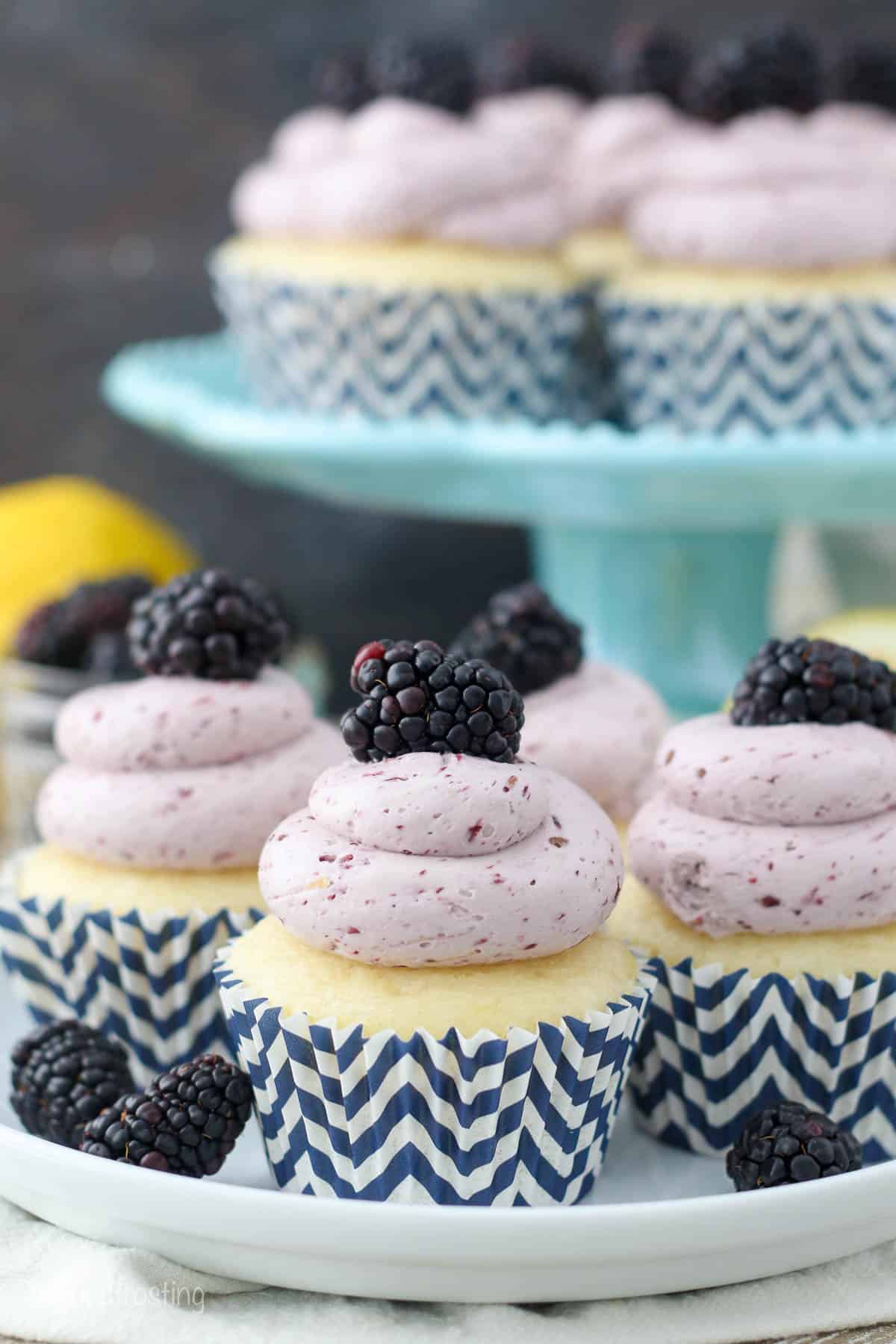 a plate of frosted lemon cupcakes topped with berries with a cake stand full of cupcakes in the background