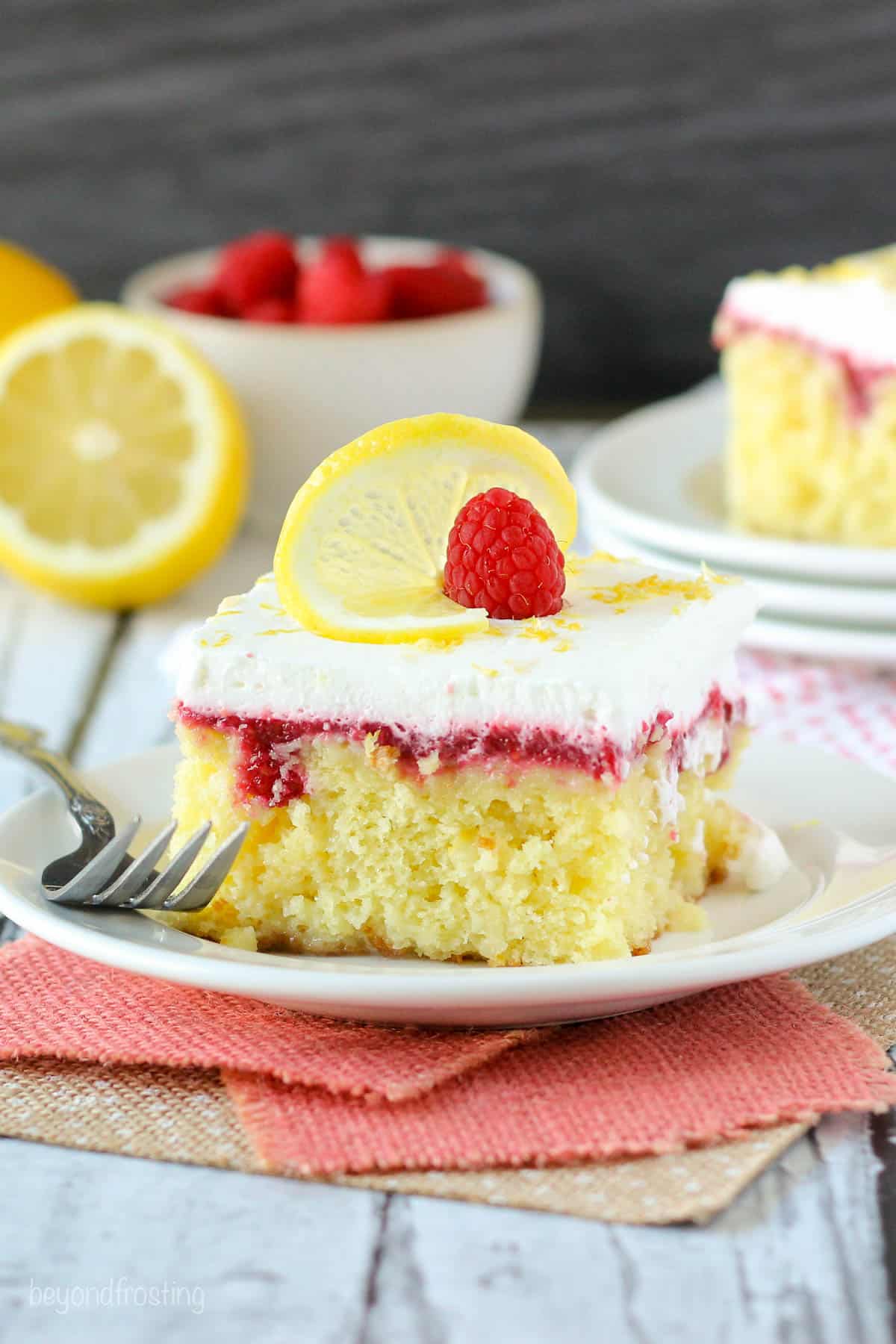 a slice of lemon cake on a plate next to a fork with a bowl of raspberries and a lemon in the background