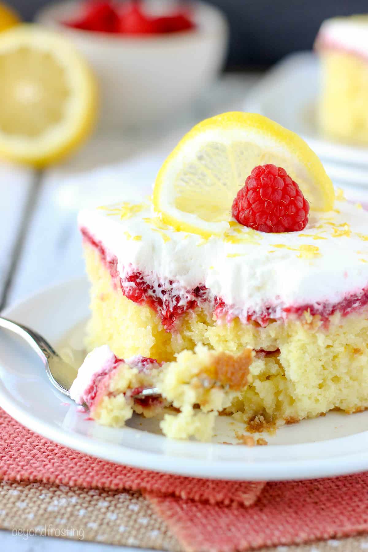 side view of a slice of lemon raspberry cake garnished with a raspberry and a lemon slice with a forkful taken out