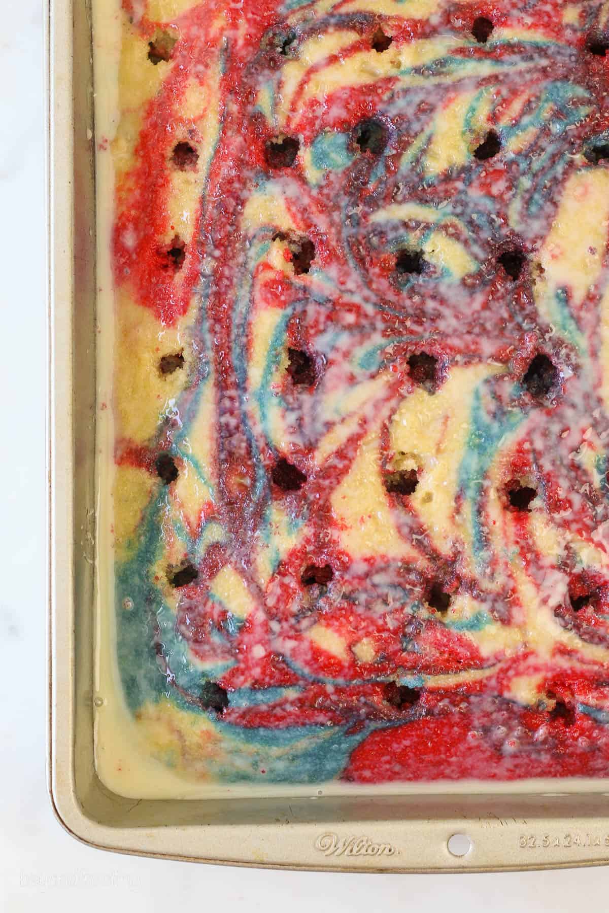 Overhead shot of a tye dye red white and blue cake with hole poked in it and sweetened condensed milk over top
