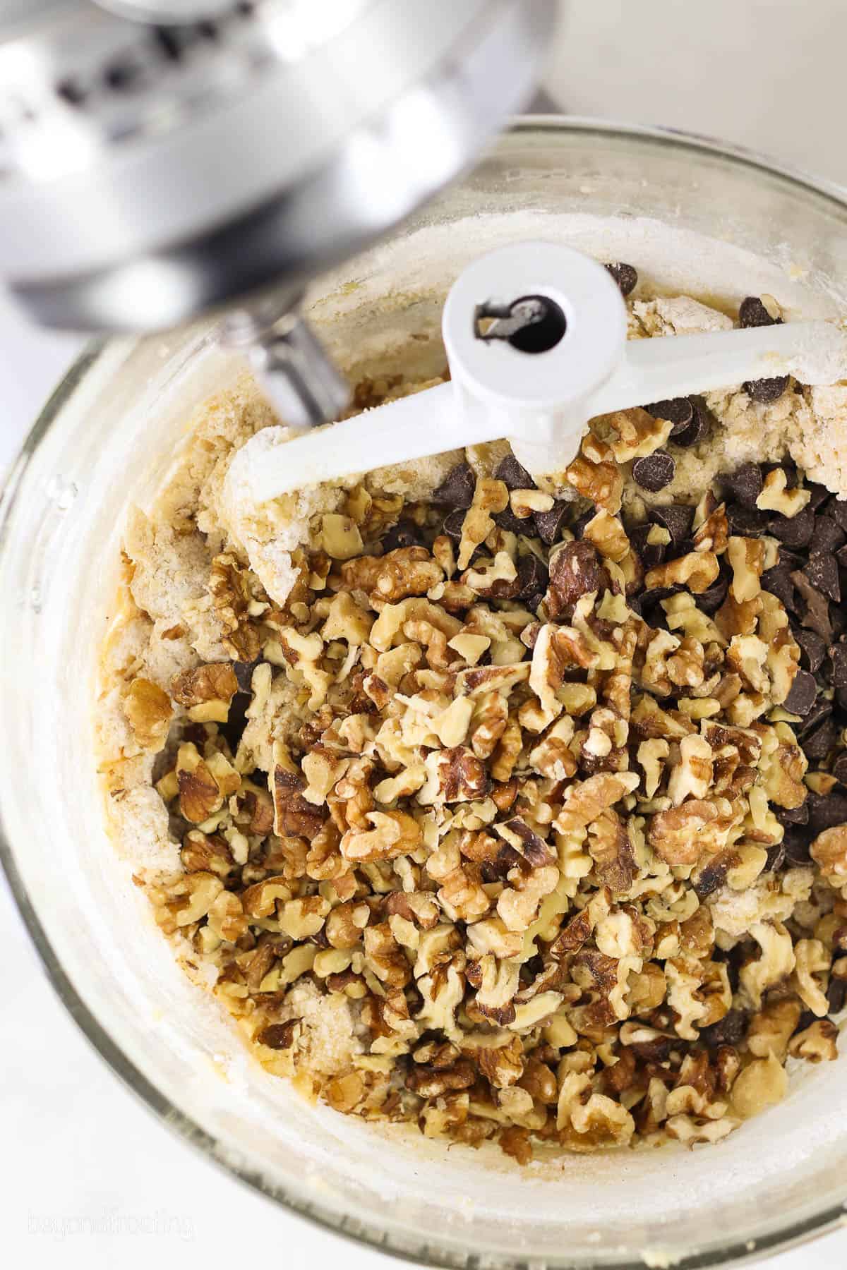A stand mixer with chopped walnuts and chocolate chips in a glass mixing bowl