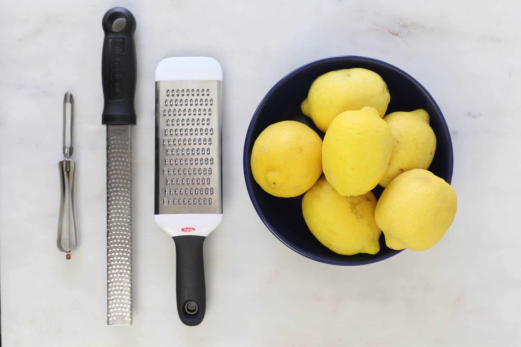 A vegetable peeler, zester, and cheese grater next to a bowl of lemons