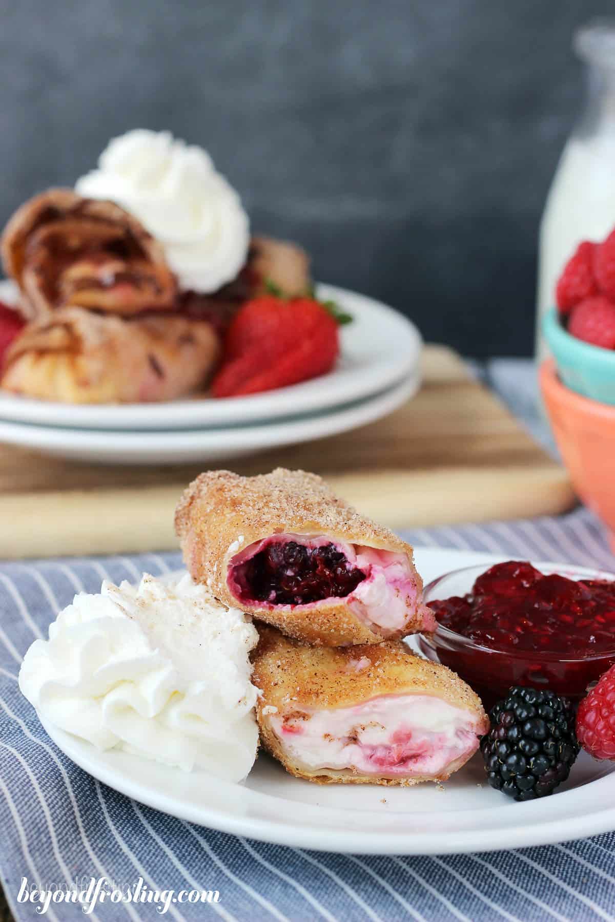 side view of two cheesecake egg rolls stacked on a plate with whip, raspberry jam, and berries