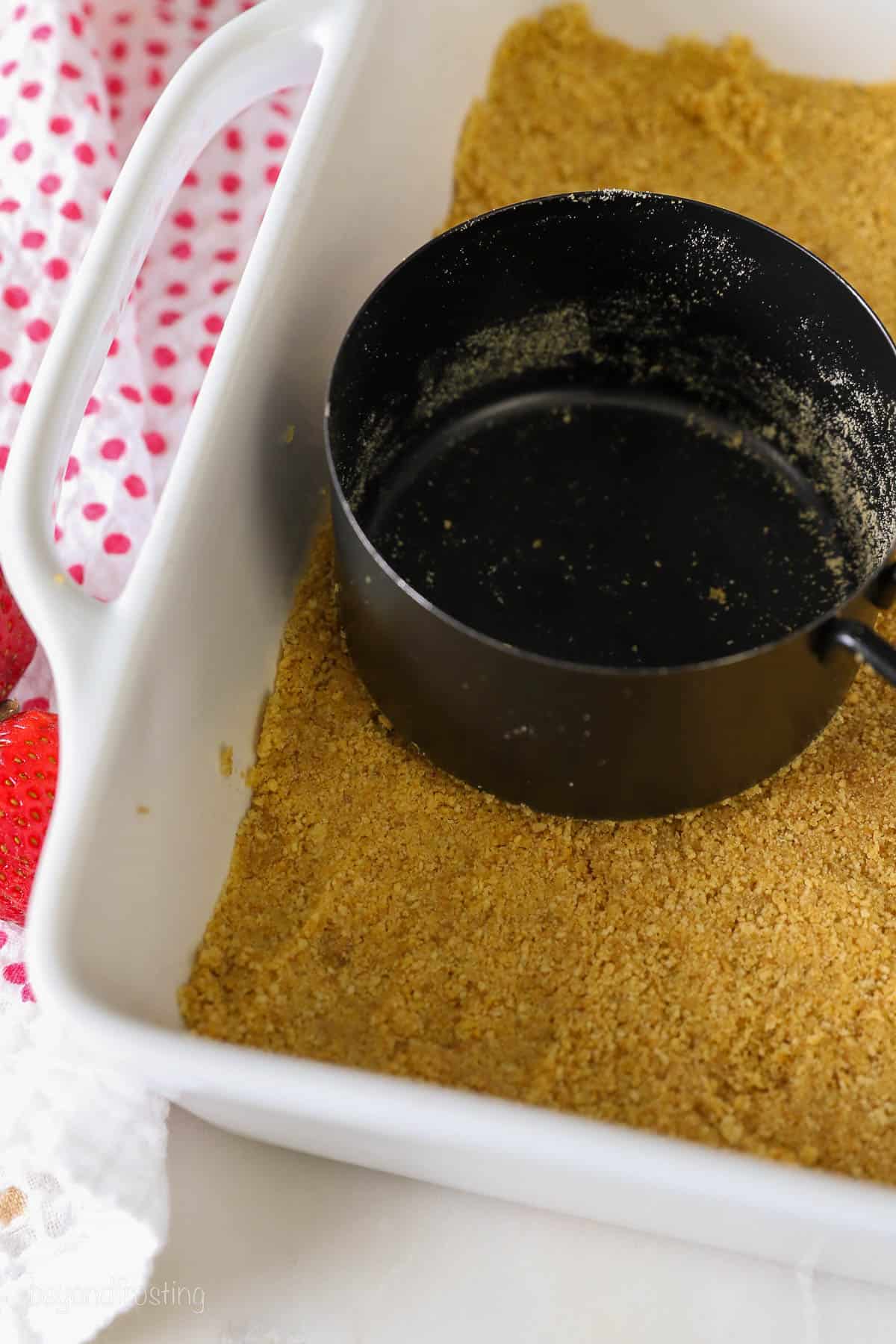 a black measuring cup pressing graham cracker crumbs into a white casserole dish