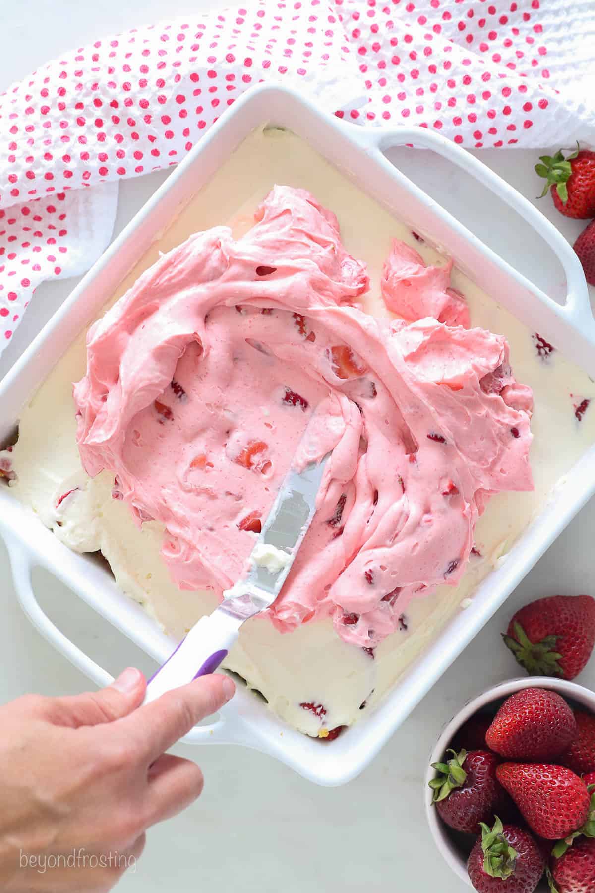 An angled spatula spreading strawberry cheesecake in a white casserole dish