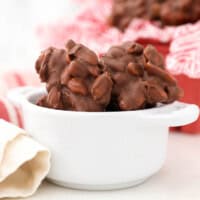 Closeup of peanut clusters in a small white bowl with more clusters in a red basket in the background.