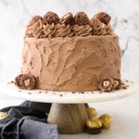 A Nutella-frosted whole Ferrero Rocher Cake sits on a white cake stand.