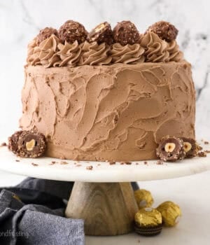 A Nutella-frosted whole Ferrero Rocher Cake sits on a white cake stand.
