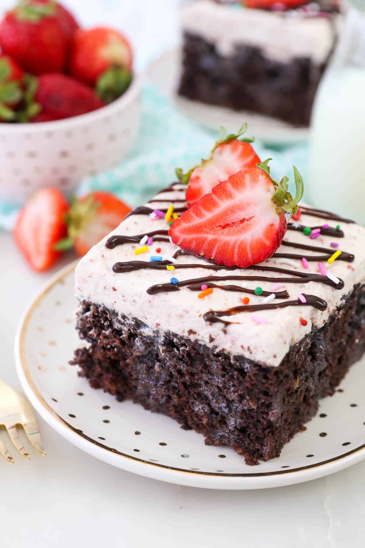 A slice of Chocolate Strawberry Milkshake Cake on a rimmed plate garnished with sprinkles, fudge and fresh strawberries