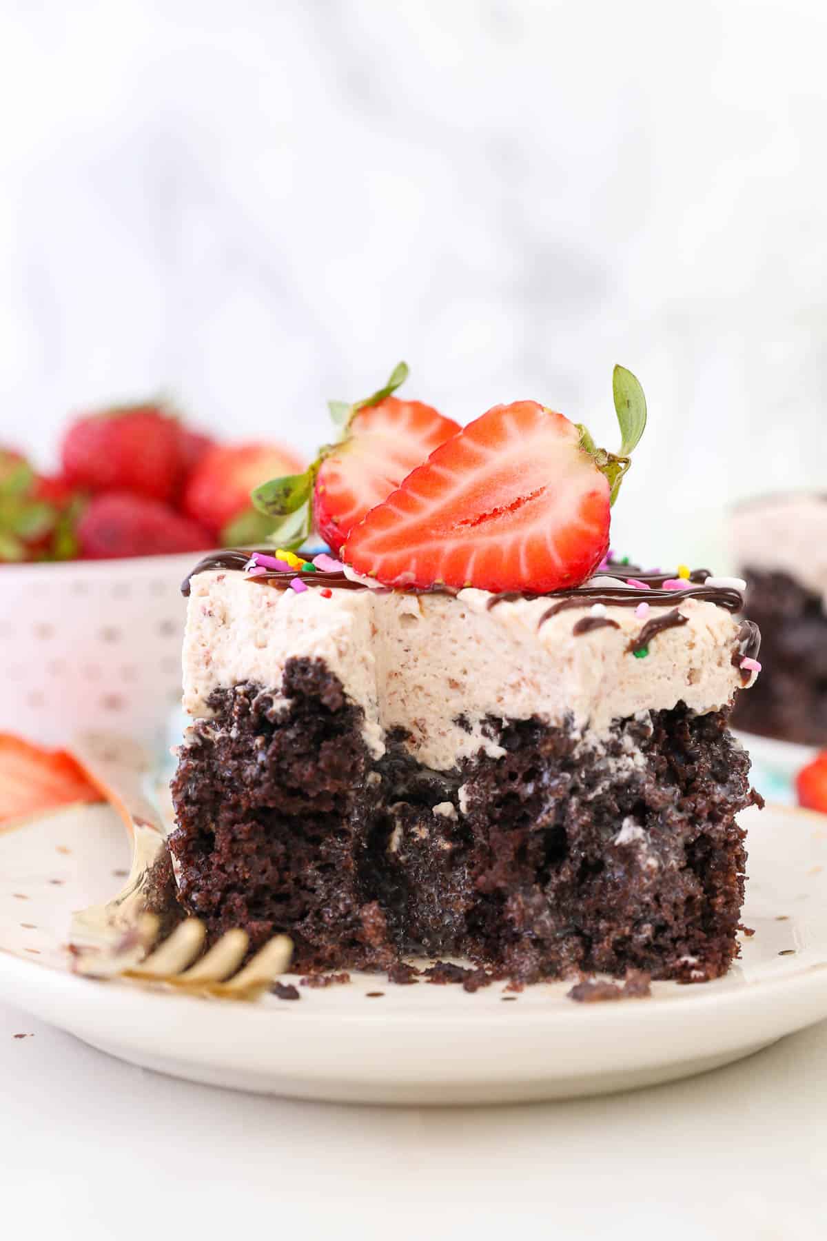 A chocolate poke cake with strawberry whipped topping, garnished with fresh strawberries, has a few minutes missing