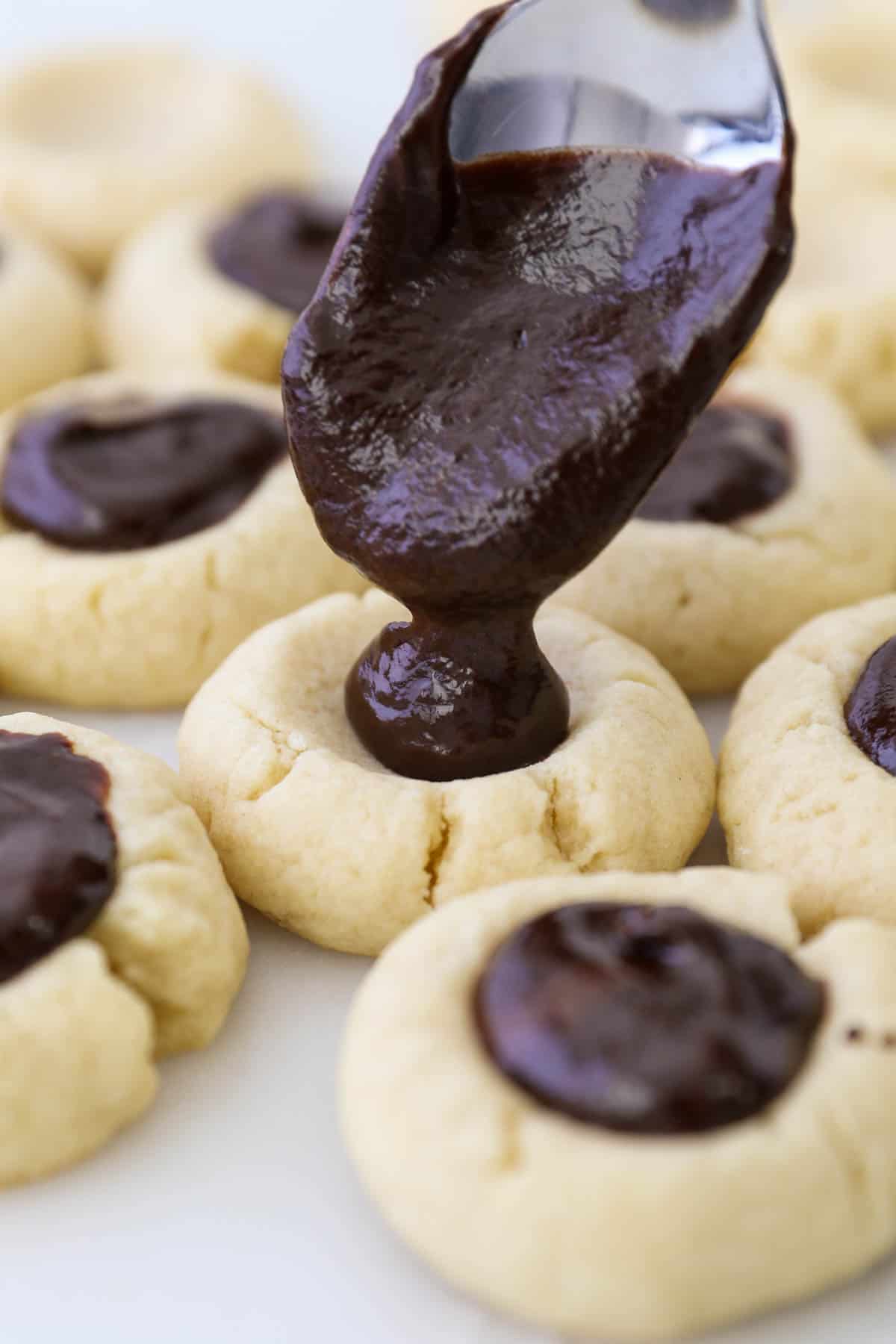 A spoon dropping chocolate ganache into the indents of baked thumbprint cookies.