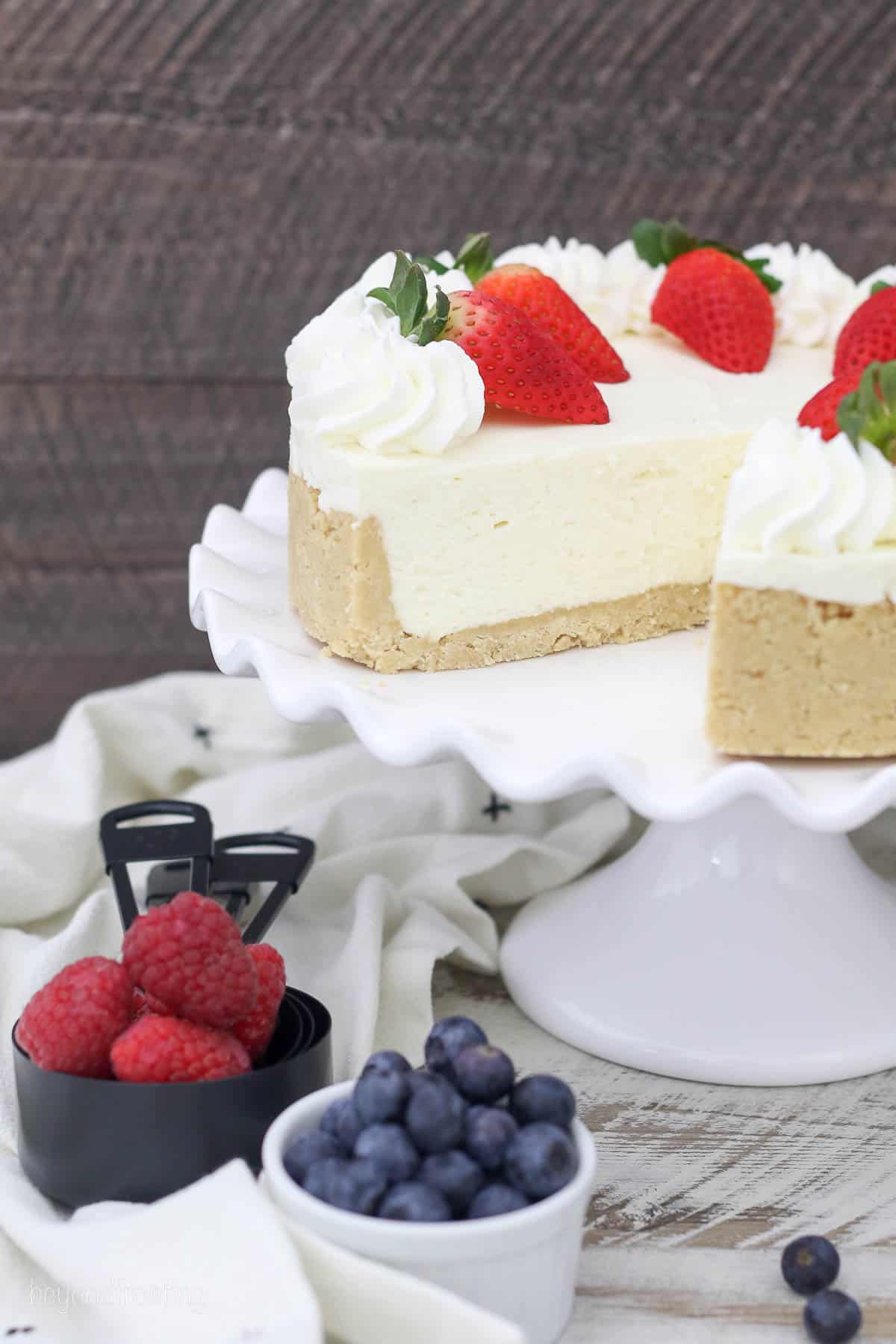 a cheesecake with a slice cut out on a cake stand surrounded by containers of fresh berries and topped with strawberries