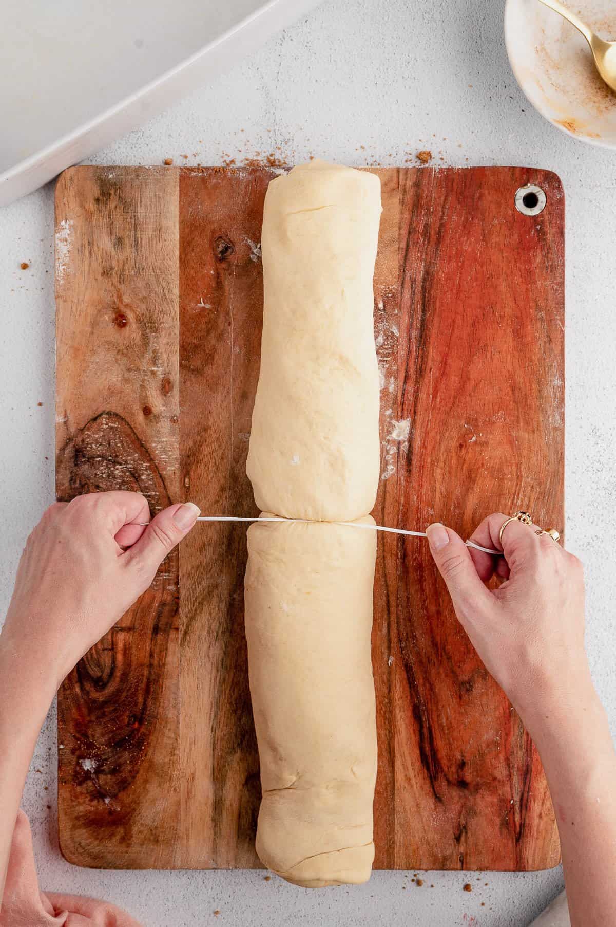 A hand uses floss to cut a log of dough into rolls on a wooden cutting board.