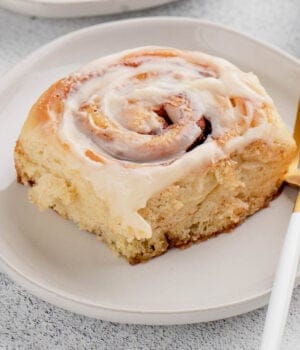 A frosted overnight cinnamon roll on a white plate next to a fork.