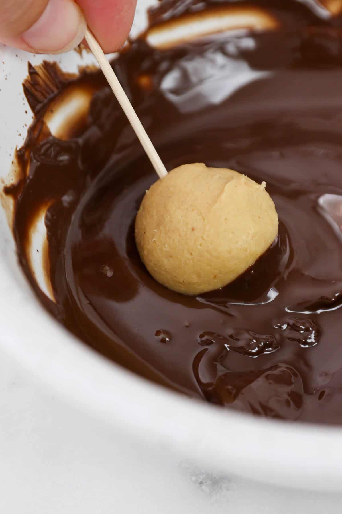 A hand dips a peanut butter ball on the end of a toothpick into a bowl of melted chocolate.