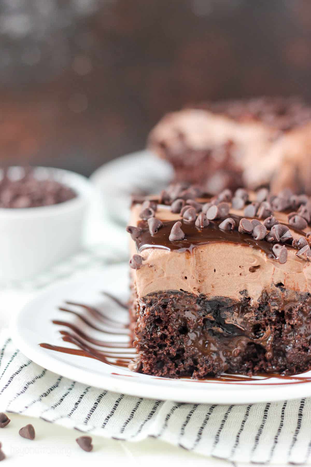 a slice of chocolate poke cake on a plate drizzled with hot fudge and topped with mini chocolate chips