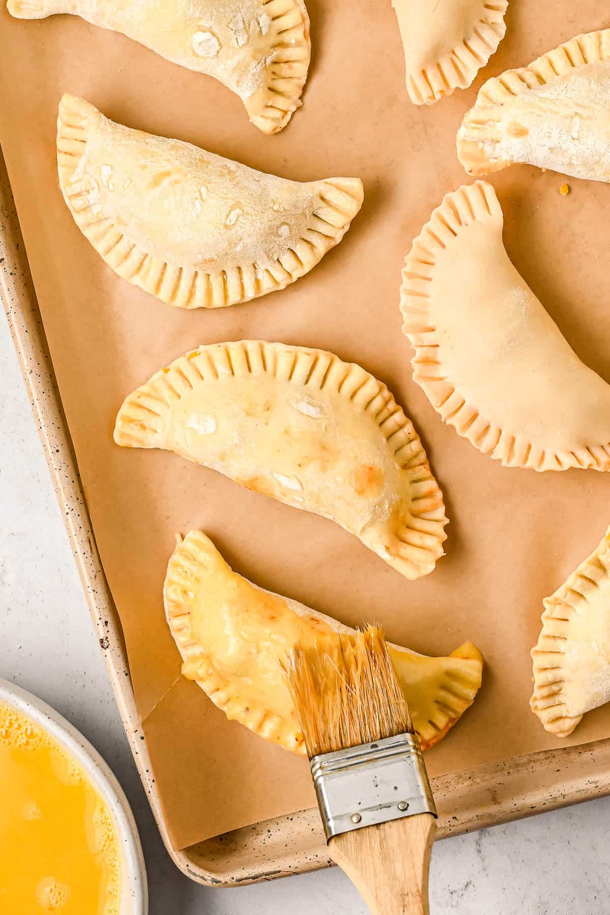 Egg wash is brushed over unbaked chicken empanadas on a baking sheet.