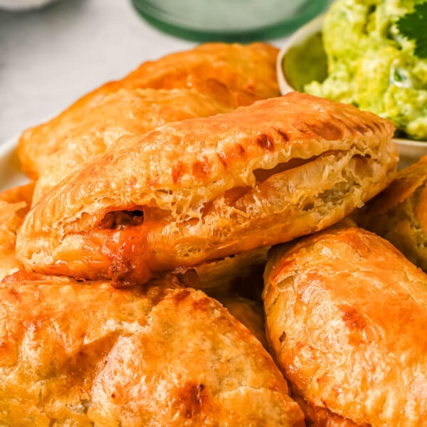 Close up of chicken empanadas stacked on a plate next to a bowl of avocado crema.