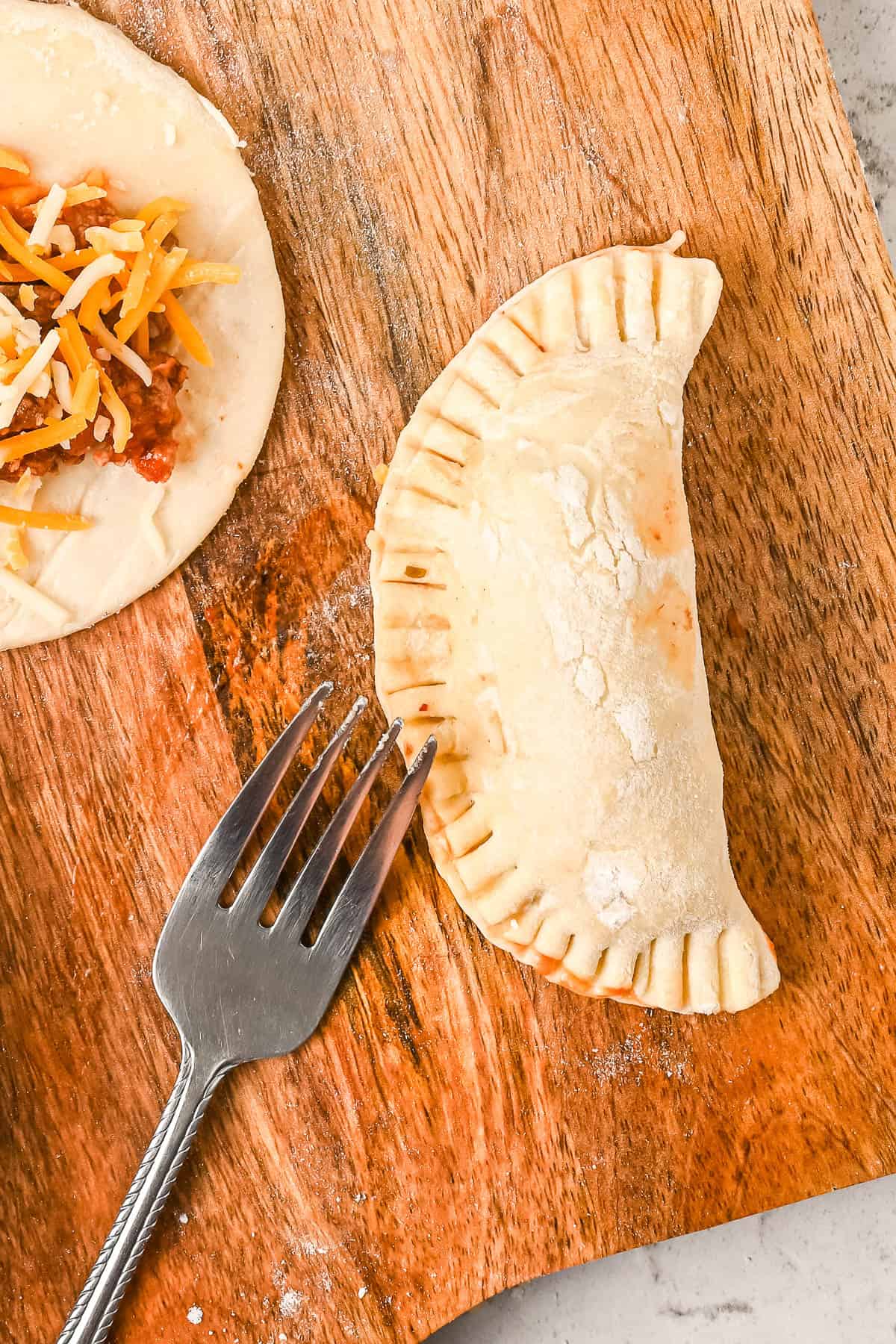 A fork is used to pinch the edges of the filled empanada dough closed.