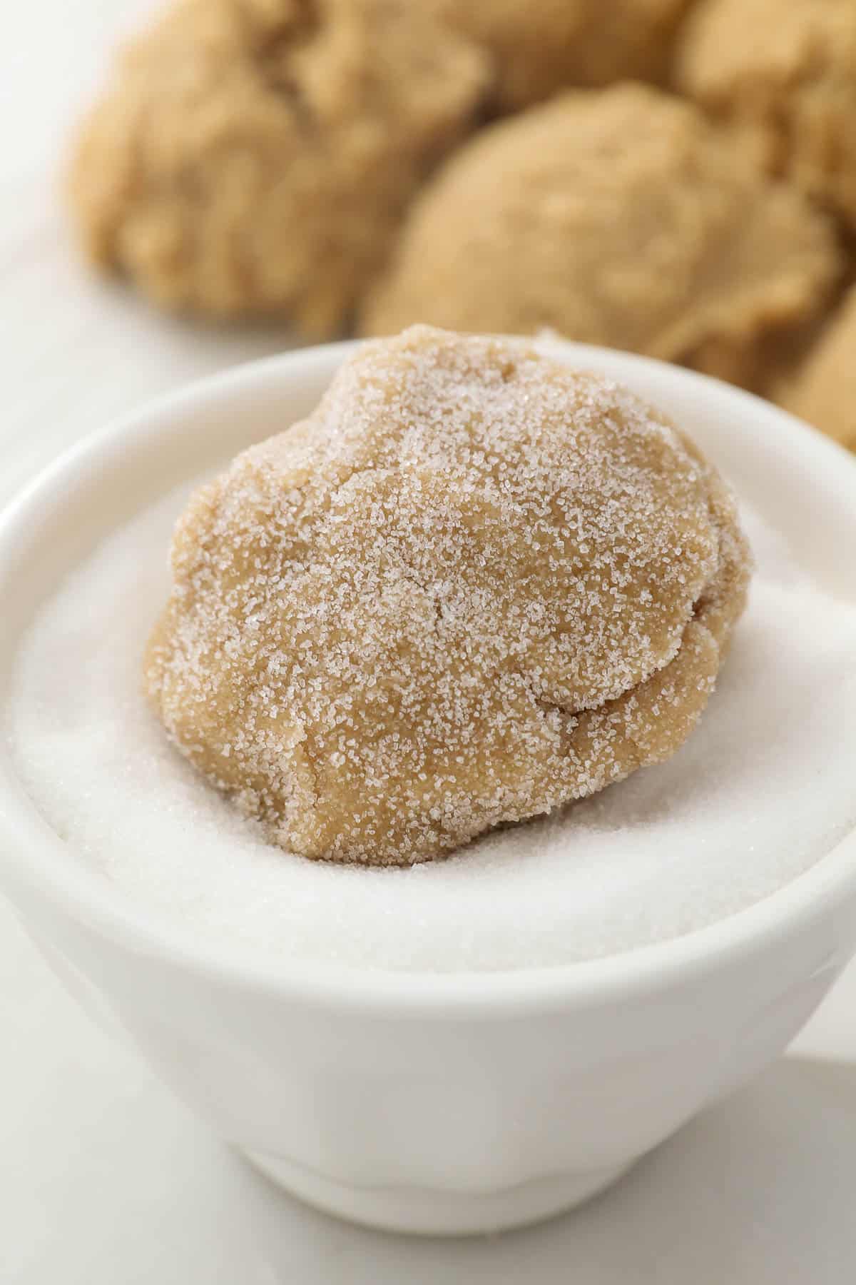 a ball of sugar cookie dough being rolled in a bowl of granulated sugar.