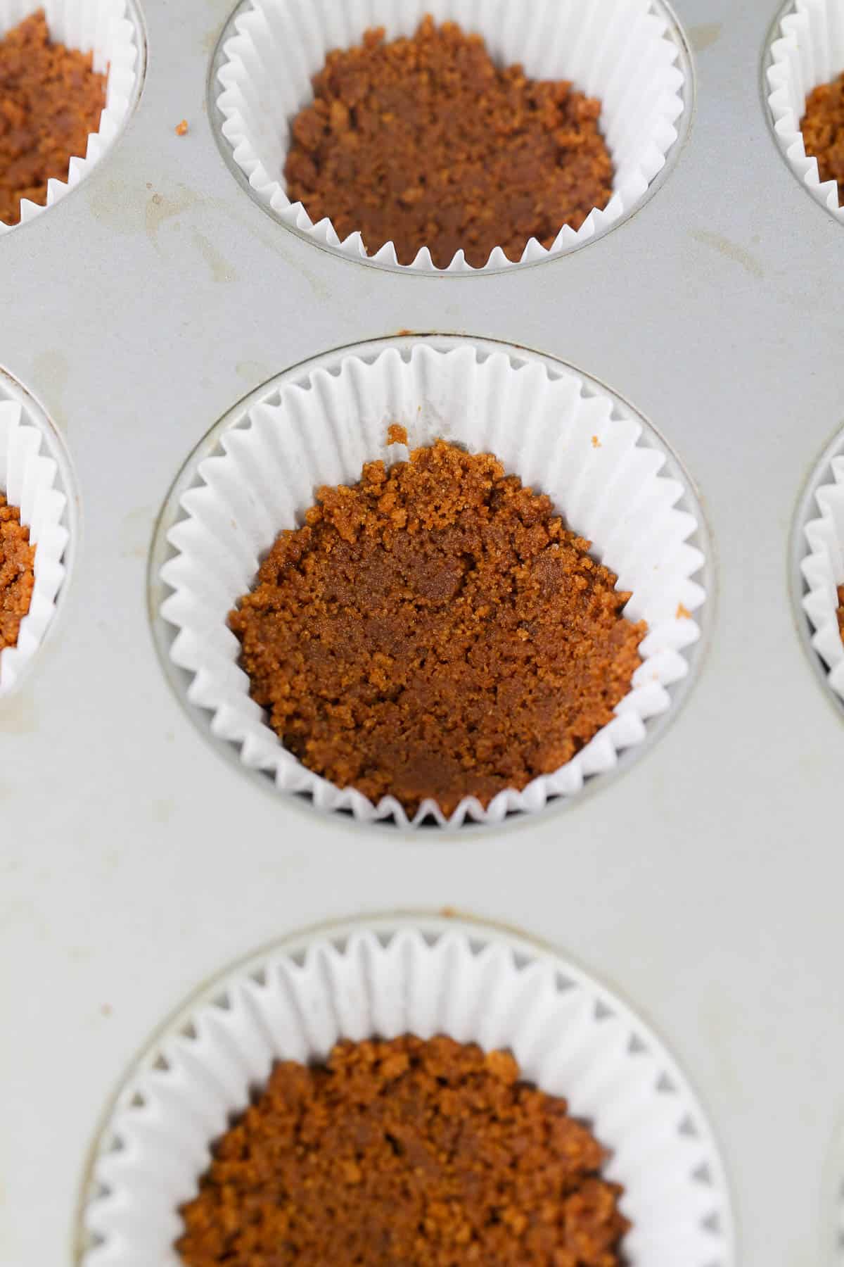 A cupcake pan with liners and a biscoff cookie crust packed in the bottom of it