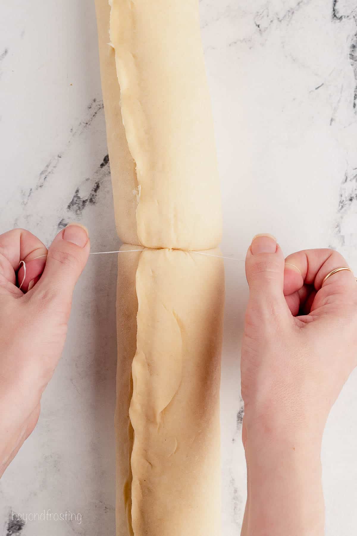 Two hands use floss to cut a log of cinnamon roll dough into rolls.