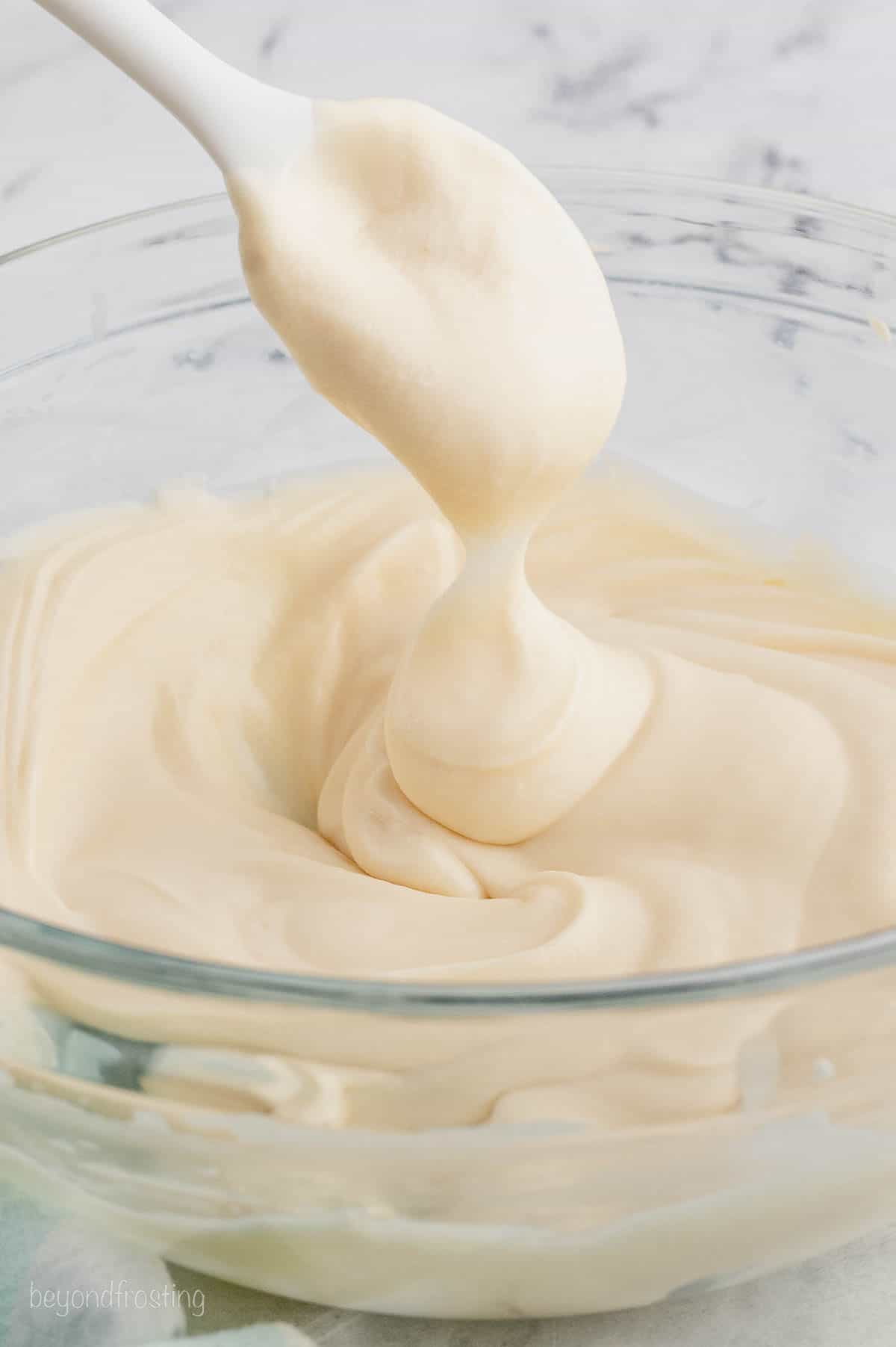 A spoon drips cream cheese frosting into a glass bowl.