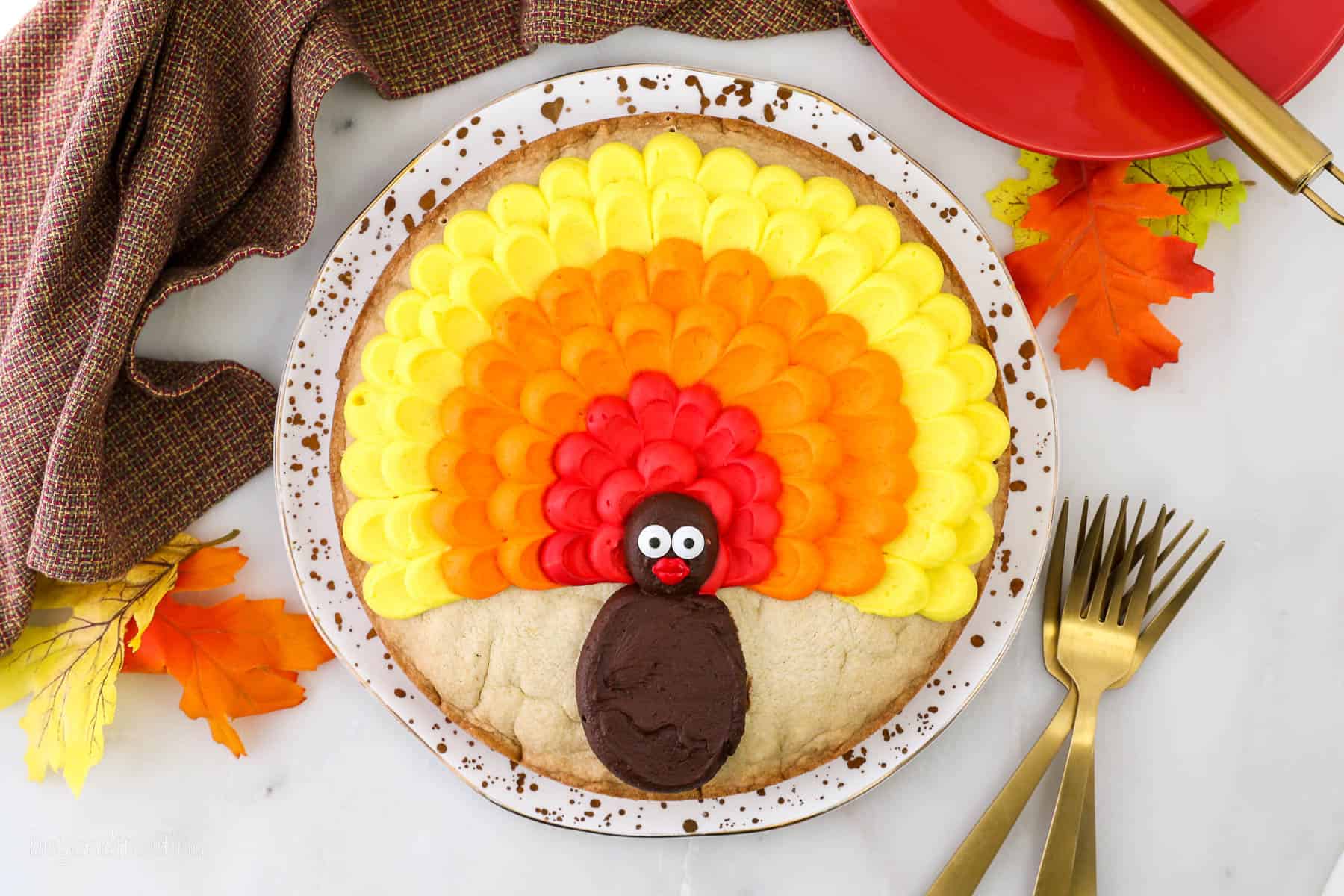 A cookie cake decorated with buttercream to look like a turkey on a plate surrounded with plates, napkin and utensils.