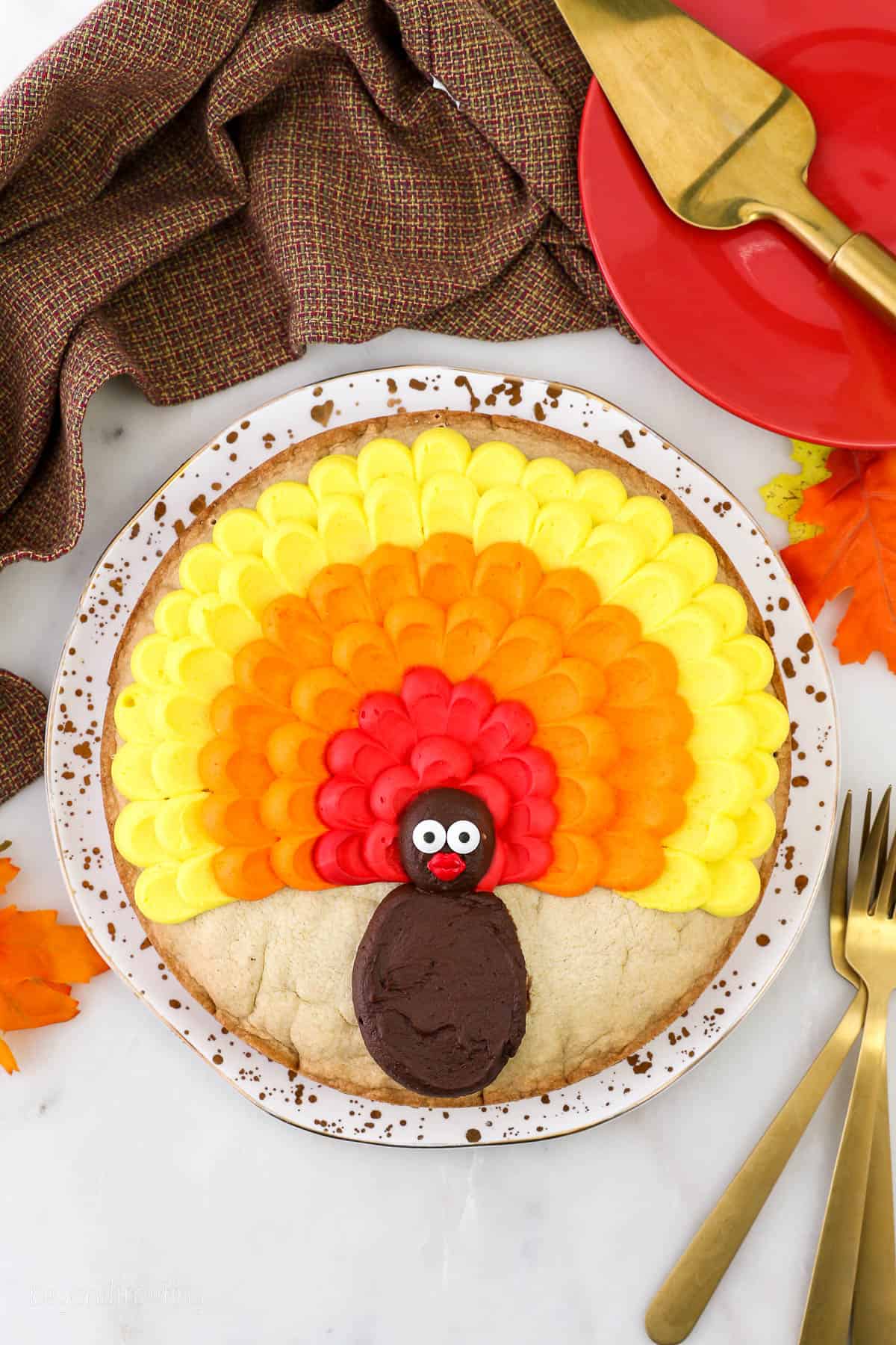 A cookie cake decorated with buttercream to look like a turkey on a plate surrounded with plates, napkin and utensils.