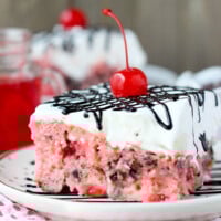 a slice of cherry poke cake on a plate topped with chocolate sauce and a cherry