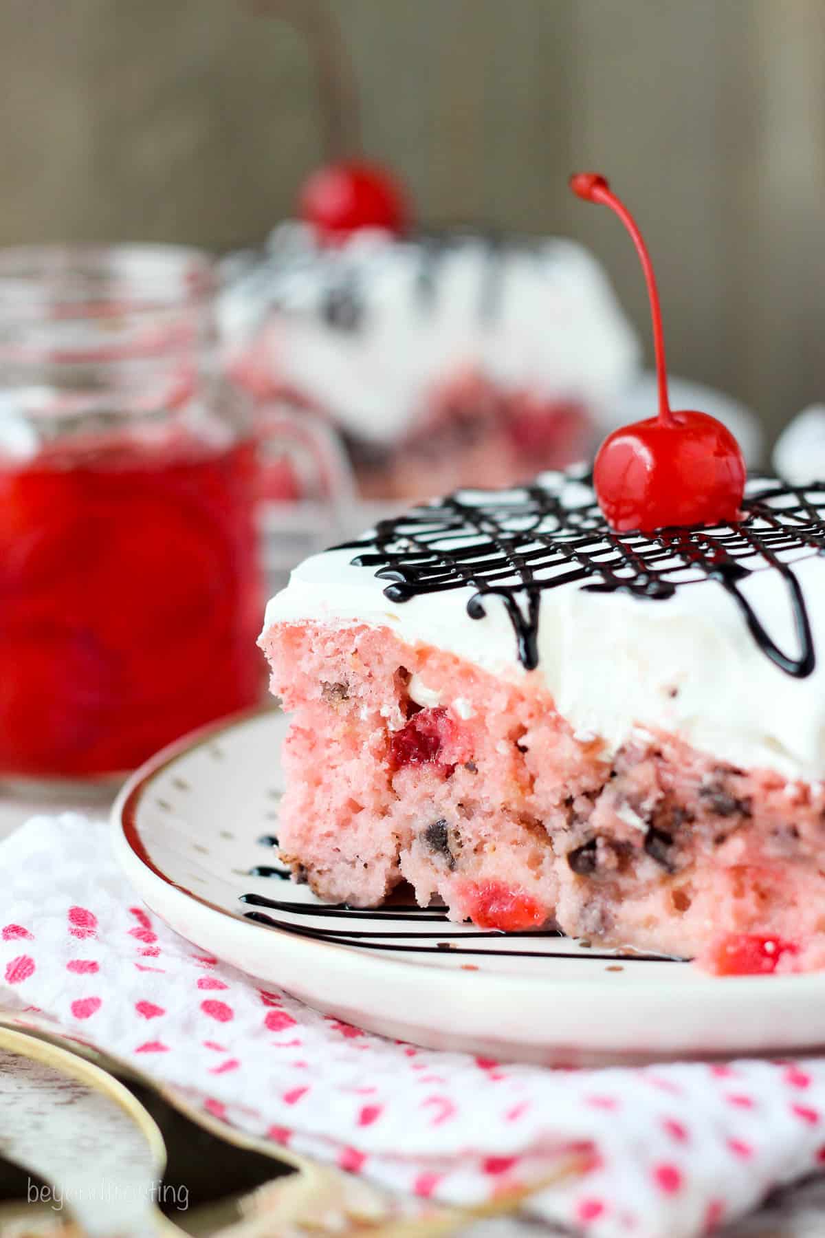 side view of a square of cherry chip poke cake with a jar of maraschino cherries in the background