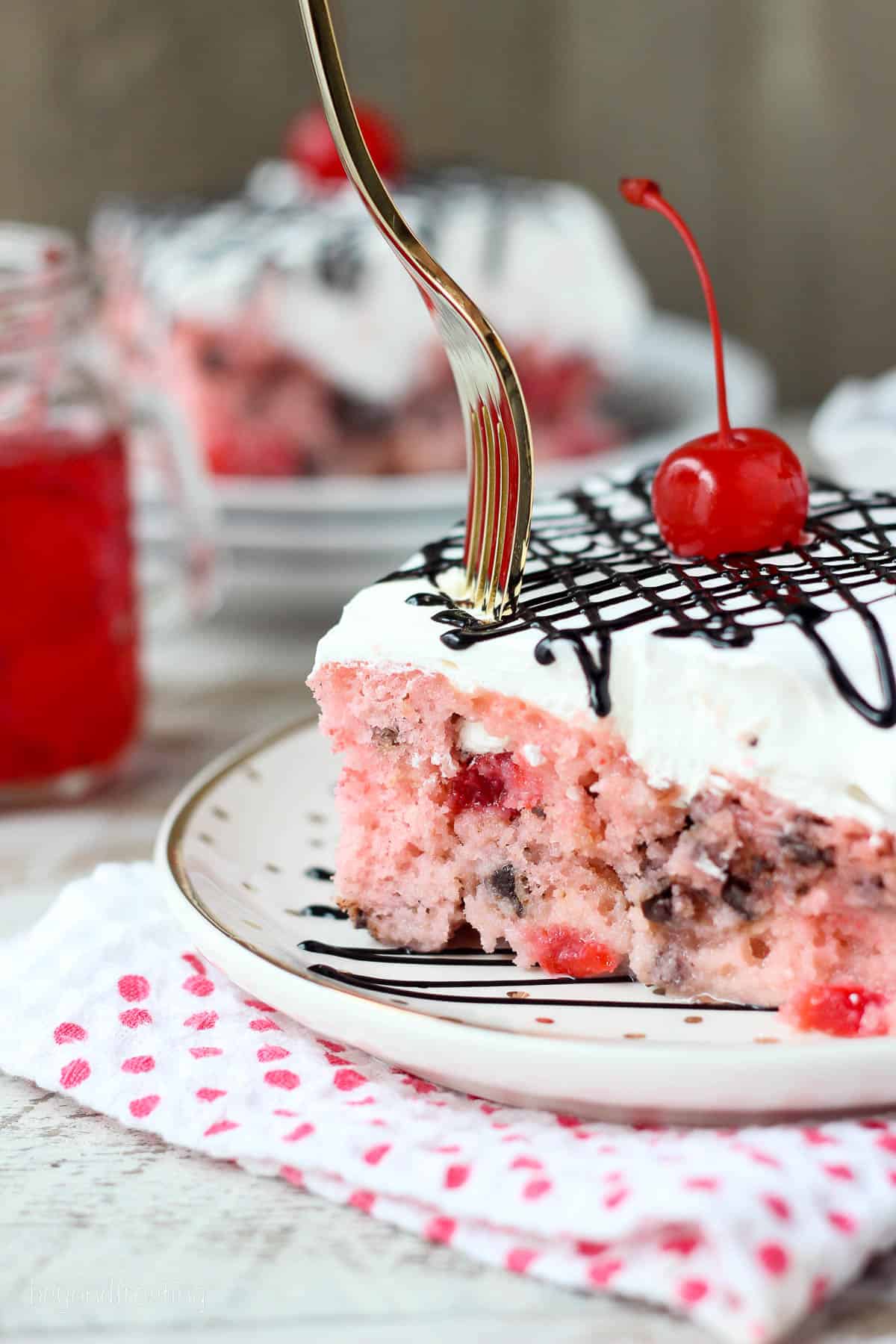 a fork being plunged into a slice of cherry poke cake topped with chocolate sauce and a maraschino cherry