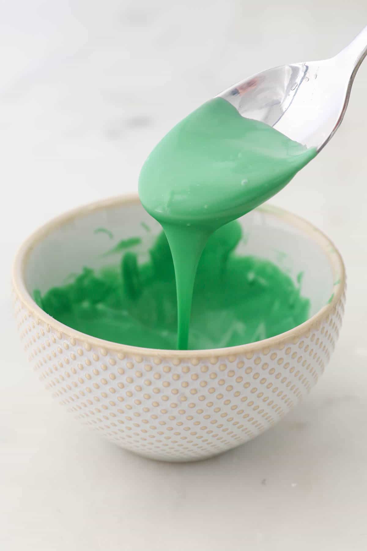 A spoon drizzles melted green candy melts into a white bowl.