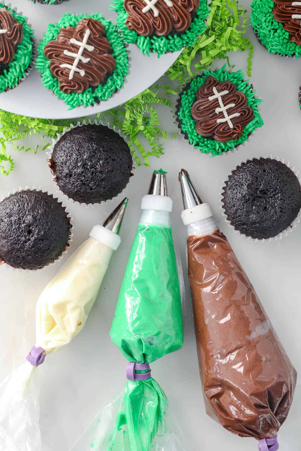 Overhead view of football cupcakes next to three piping bags fitted with tips, filled with white, green, and brown buttercream frosting.