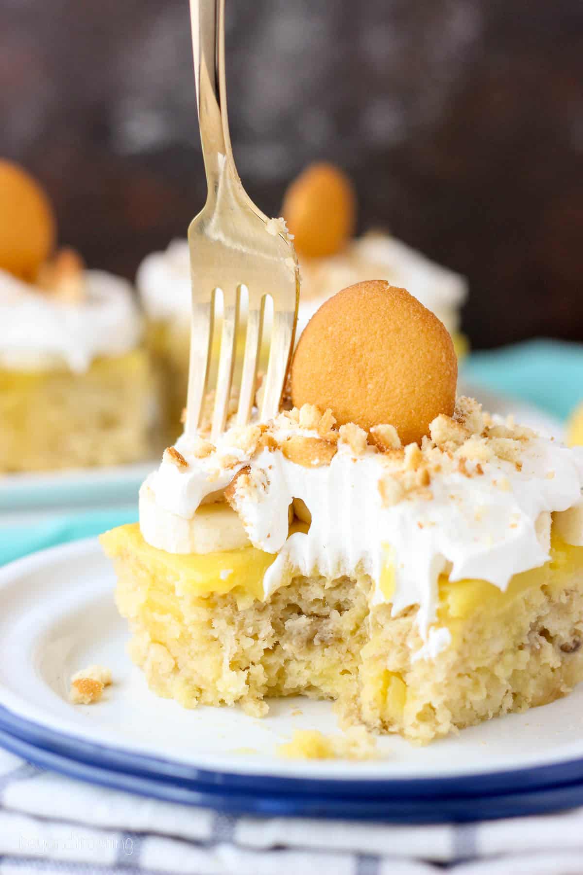 a fork being plunged into a slice of banana poke cake topped with a nilla wafer.