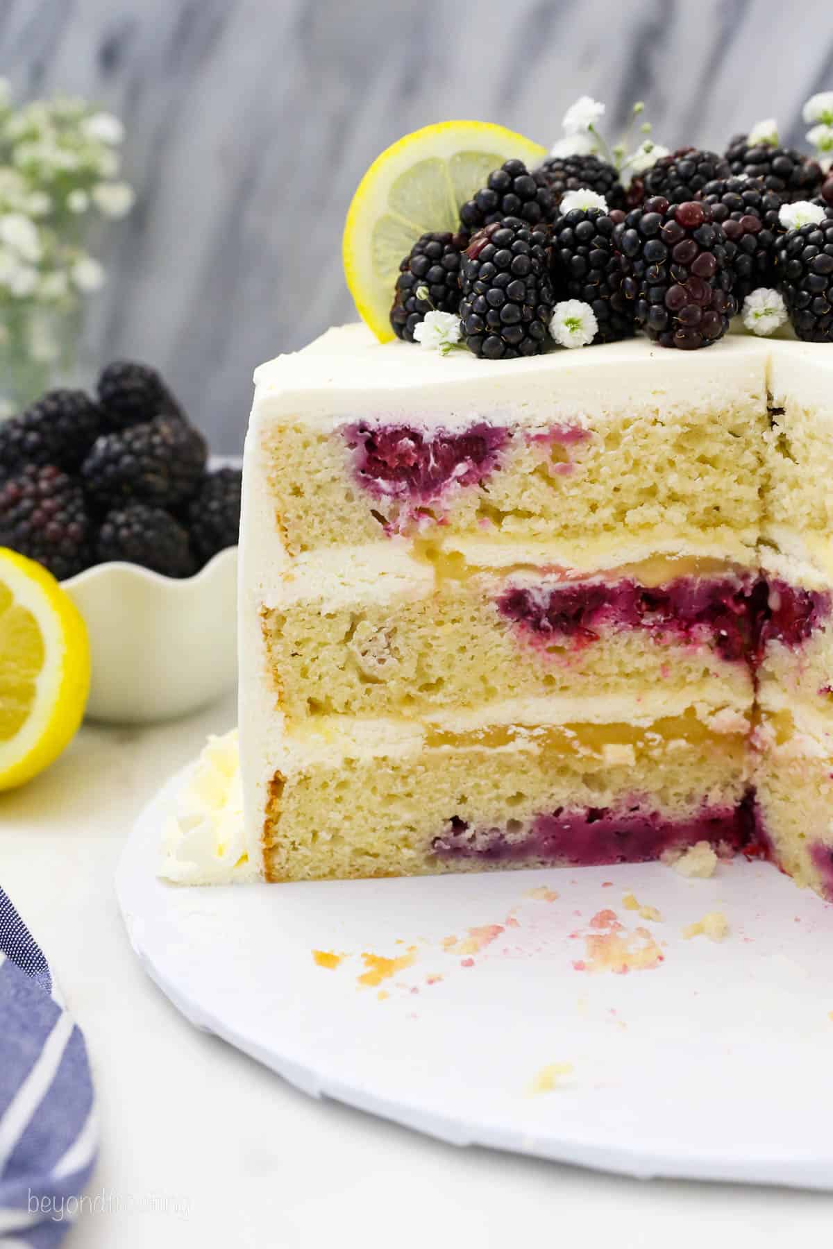 side view of a lemon cake with a slice cut out topped with fresh blackberries and lemon.