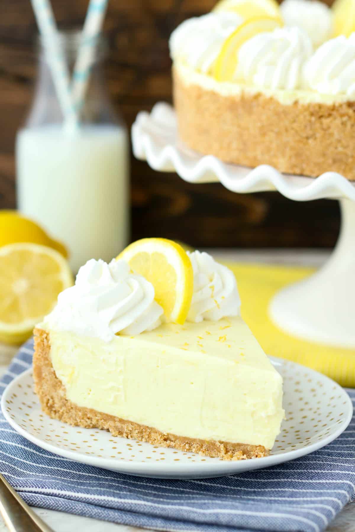 side view of a slice of lemon pie on a plate next to a cake stand with the whole pie