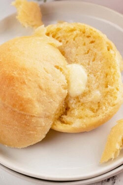 Two halves of a dinner roll on a white plate, with a pat of butter melting on one half.
