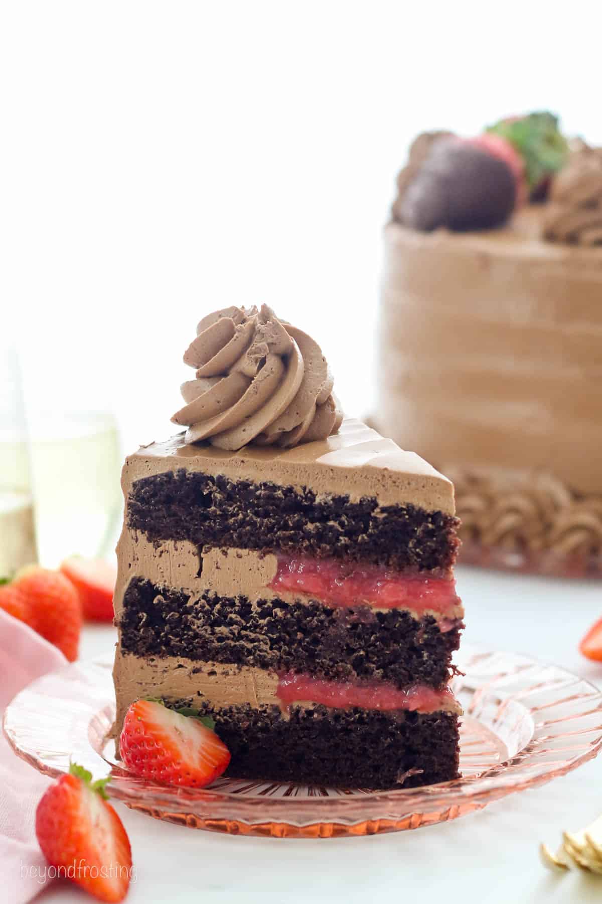 An upright slice of chocolate strawberry cake topped with a chocolate frosting swirl on a plate, with the rest of the cake in the background.