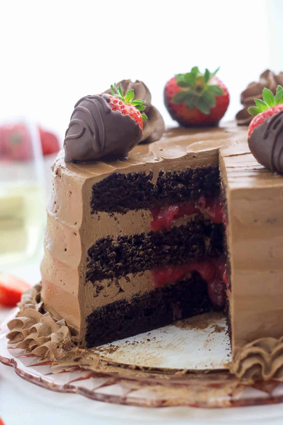 Chocolate strawberry cake decorated with chocolate frosting swirls and chocolate covered strawberries, with a slice missing to show the strawberry filled layers inside.