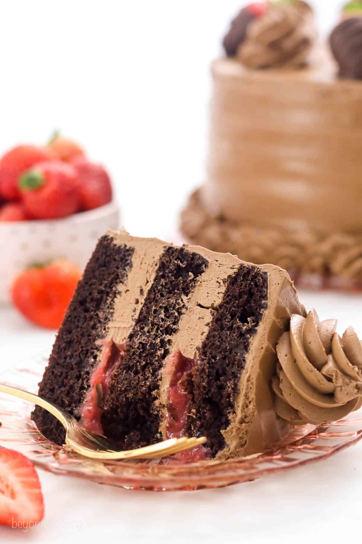 A partially eaten slice of frosted chocolate strawberry layer cake laying on its side on a plate, next to a fork.