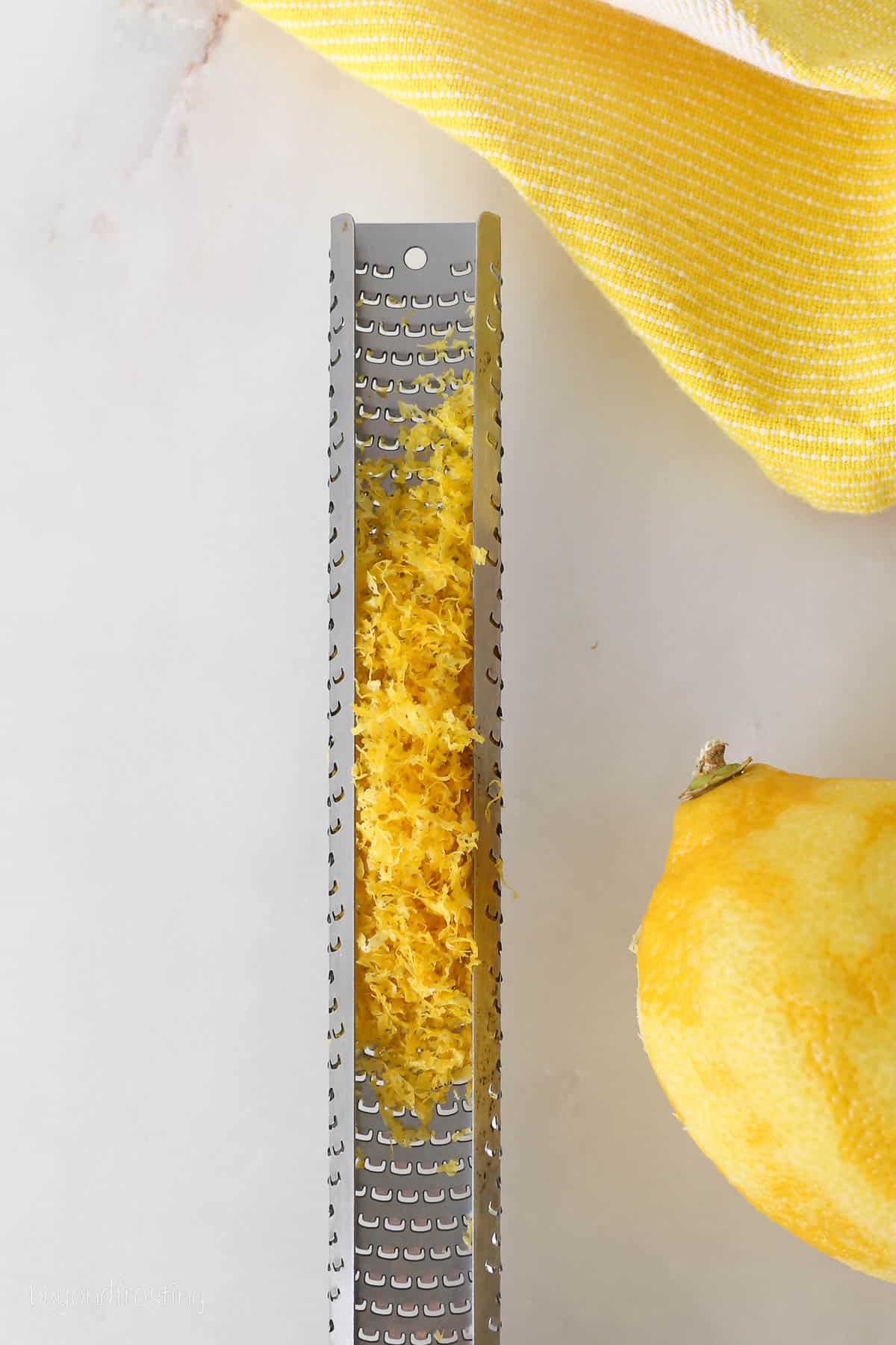 A lemon zester with zest, next to a zested lemon.